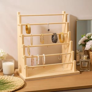 May include: A light-wood jewelry display stand with several horizontal rods for hanging bracelets, necklaces, and earrings. The stand holds various gold and beaded jewelry. A lit candle and a mirror are in the background. The stand is on a wooden surface.