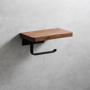 Puede incluir: Un accesorio de baño moderno con una estantería de madera y un portarrollos de papel higiénico de metal negro. La estantería es de madera marrón claro, y el soporte tiene un diseño curvo. El conjunto está montado en una pared gris claro.