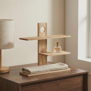 May include: A three-tiered wooden shelf unit with a circular cutout and a perfume bottle. The unit is made of light-colored wood and has a rectangular base. A folded, light-colored cloth rests on the base.
