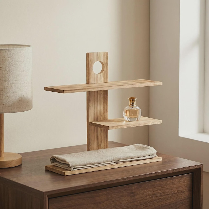 May include: A three-tiered wooden shelf unit with a circular cutout and a perfume bottle. The unit is made of light-colored wood and has a rectangular base. A folded, light-colored cloth rests on the base.