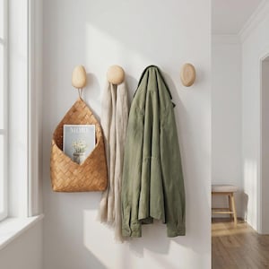Puede incluir: Una solución de almacenamiento montada en la pared con una cesta tejida, ganchos de madera y una chaqueta verde. La cesta contiene una revista con la palabra "MORE" visible. Una bufanda beige cuelga de un gancho. Los ganchos son de madera clara.