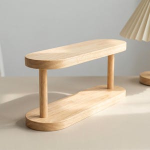 May include: A light-colored wooden display stand with an oval top and base, connected by two cylindrical supports. The stand is made of natural wood and has a minimalist design, suitable for displaying small items.