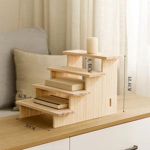 May include: A wooden display stand with three steps, 11.8 inches tall, 11.8 inches wide, and 2.4 inches deep. The natural wood-colored stand holds a small, cylindrical vase and two books. The stand is designed to showcase small items.