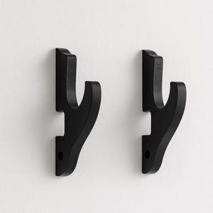 Könnte beinhalten: Zwei schwarze Wandhaken mit geschwungenem Design. Jeder Haken hat eine flache Basis mit einem einzelnen Loch zur Befestigung und einen gebogenen Arm. Die Haken bestehen aus einem soliden Material und sind zum Aufhängen von Gegenständen bestimmt.
