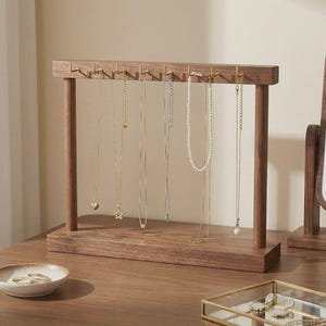 May include: A wooden jewelry display stand with multiple necklaces hanging from it. The stand is made of dark wood and has a rectangular base with two vertical supports. The necklaces are of varying lengths and styles, including gold and pearl necklaces.