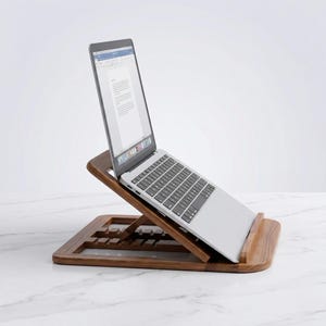 May include: A wooden laptop stand with a laptop on top. The stand is made of dark brown wood and has a sloped design. The laptop is silver and has a black keyboard. The screen displays a document.