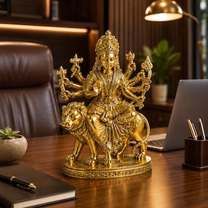 May include: A metallic gold-coloured statue of a multi-armed deity seated atop a lion. The figure wears an ornate crown and traditional jewellery, with hands positioned in various mudras and holding symbolic items, all set on a decorative base.