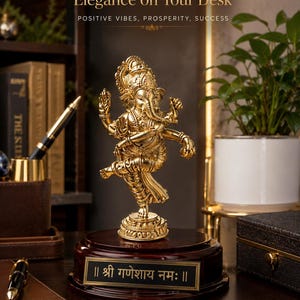 May include: A gold-colored statue of the Hindu deity Ganesha, mounted on a dark wooden base with a gold plaque. The plaque features text in a foreign language. The statue is in a dancing pose. The image includes the text "Elegance on Your Desk" and "POSITIVE VIBES, PROSPERITY, SUCCESS".