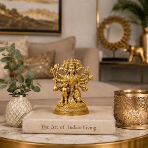 K&ouml;nnte beinhalten: Eine goldfarbene Statue einer Gottheit mit mehreren Armen und Gesichtern, die auf einem dekorativen Sockel steht. Die Statue steht auf einem Tisch mit einem Buch mit der Aufschrift "The Art of Indian Living". Ein goldfarbener Kerzenhalter und eine Vase mit Gr&uuml;nzeug stehen ebenfalls auf dem Tisch.