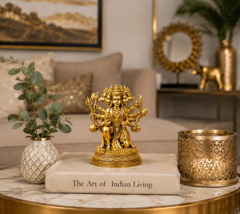 K&ouml;nnte beinhalten: Eine goldfarbene Statue einer Gottheit mit mehreren Armen und Gesichtern, die auf einem dekorativen Sockel steht. Die Statue steht auf einem Tisch mit einem Buch mit der Aufschrift "The Art of Indian Living". Ein goldfarbener Kerzenhalter und eine Vase mit Gr&uuml;nzeug stehen ebenfalls auf dem Tisch.