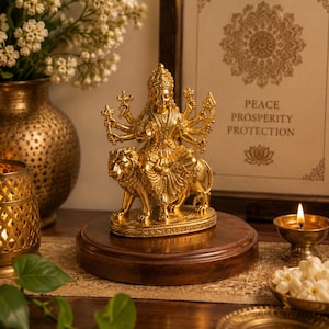 Könnte beinhalten: Eine goldfarbene Statue einer Gottheit, die auf einem Löwen reitet, auf einem Holzsockel. Die Statue hat mehrere Arme und ist sehr detailliert. Der Hintergrund enthält einen gerahmten Druck mit den Worten "PEACE PROSPERITY PROTECTION".