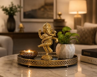 May include: A gold-coloured statue of Ganesha, the Hindu deity, dancing on a decorative tray. The tray also holds a small potted plant and a lit candle in a gold bowl. The items are arranged on a marble-topped table.