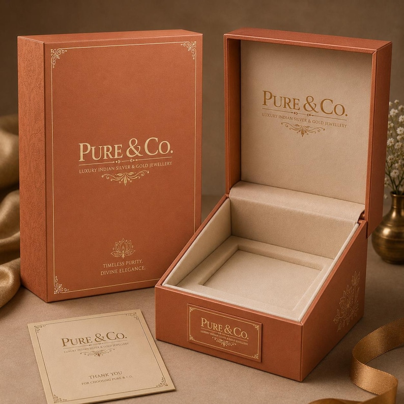 K&ouml;nnte beinhalten: Ge&ouml;ffnete orangefarbene Schmuckschatulle mit beigem Innenraum und dem Text "Pure & Co." in goldfarbenen Buchstaben. Eine passende Schachtel befindet sich dahinter, und eine kleine Karte mit demselben Logo davor. Die Schachteln sind f&uuml;r luxuri&ouml;sen indischen Silber- und Goldschmuck.