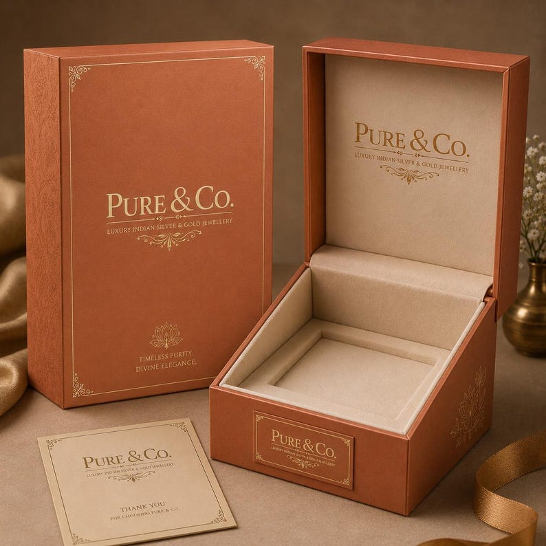 May include: Open orange jewelry box with a soft beige interior and the text "Pure & Co." in gold lettering. A matching box is closed in the background, and a small card with the same branding is in the foreground. The box is designed for luxury Indian silver and gold jewelry.