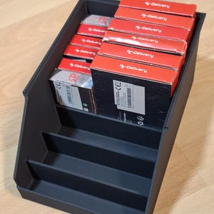 Könnte beinhalten: Ein schwarzer, abgestufter Aufbewahrungsorganizer mit mehreren rechteckigen Schachteln. Die Schachteln sind überwiegend rot und tragen den weißen Schriftzug "A-Delivery" auf der Oberkante. Einige Schachteln zeigen Etiketten mit Barcodes.
