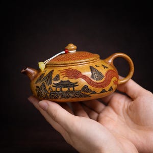 Pode incluir: Pequena chaleira ornamentada, em tons de castanho e laranja, com um design detalhado de dragão em vermelho. A chaleira tem uma alça curva, um bico e uma tampa com um botão decorativo. O design inclui uma paisagem com montanhas e uma pagoda.