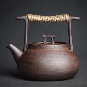 Könnte beinhalten: Eine braune Keramik-Teekanne mit einem Seilgriff. Die Teekanne hat einen Ausguss und einen Deckel mit einem kleinen Griff. Der Griff besteht aus einem dicken Seil, das um einen Metallrahmen gewickelt ist. Die Teekanne ist dunkelbraun und hat eine strukturierte Oberfläche.