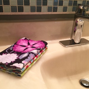 May include: A stack of four colorful hand towels with a floral pattern. The towels are folded and placed on a white bathroom sink counter.