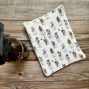 May include: White rectangular cloth featuring a pattern of mason jars filled with flowers in yellow, red, and green. The cloth has a cream-coloured fringe. The background is a wooden surface.