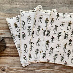May include: Four white square cloths with a repeating pattern of black line-drawn mason jars filled with various flowers. The cloths have a cream-coloured, fringed edge. A brown glass bottle with a pump is visible on the left side of the image.