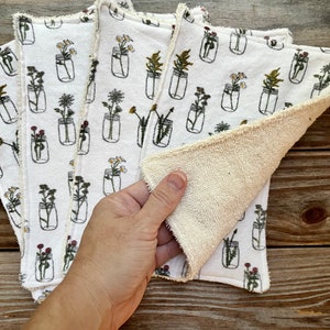 May include: White cloth napkins featuring a floral mason jar design. The napkins have a cream-coloured, textured reverse side and a decorative edge. The floral design includes various flowers in black outline jars.