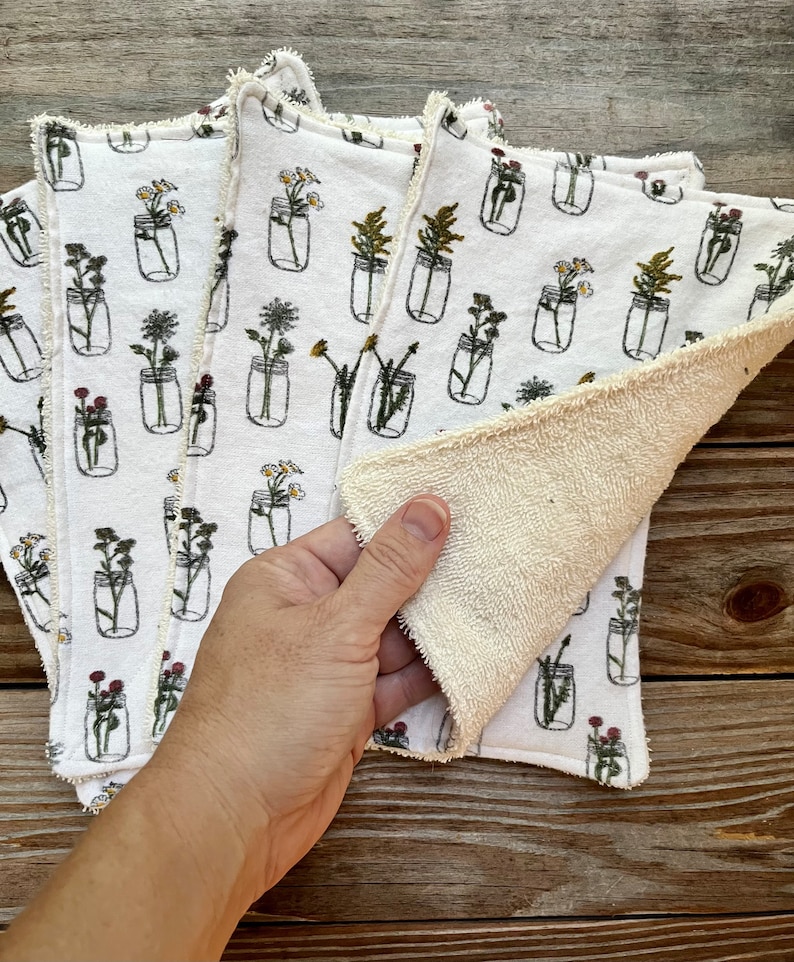 May include: White cloth napkins featuring a floral mason jar design. The napkins have a cream-coloured, textured reverse side and a decorative edge. The floral design includes various flowers in black outline jars.