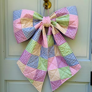 Peut inclure: Un grand nœud papillon en vichy coloré, avec un motif patchwork de carrés roses, verts, bleus et beiges. Le nœud est suspendu à une porte bleu clair et est noué au centre avec une boucle pour l'accrocher.