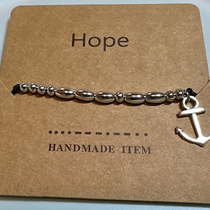 May include: A silver beaded bracelet with an anchor charm, displayed on a brown card. The card reads "Hope" at the top and "HANDMADE ITEM" at the bottom. The bracelet features oval and round beads.