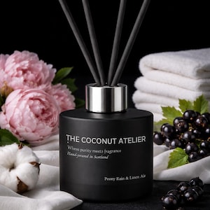 May include: A black diffuser bottle with a silver collar and black reeds. The bottle has text that reads "THE COCONUT ATELIER" and "Peony Rain & Linen Air". The background includes pink peonies, white towels, and black berries.