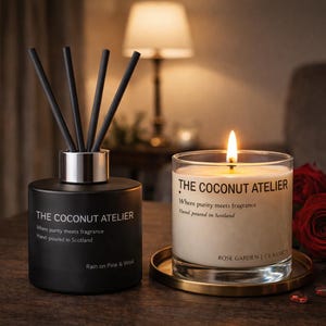 May include: A black diffuser and a lit candle from The Coconut Atelier. The candle is in a glass jar on a gold-coloured coaster, with red roses and white flowers in the background. The diffuser has black reeds.