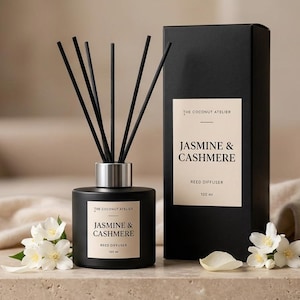 May include: A black Jasmine & Cashmere reed diffuser with black reeds and a silver collar, next to a matching box. The product is displayed with white flowers and the words "Soft & Elegant".