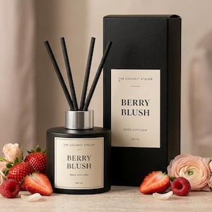 May include: A black reed diffuser with the text "Berry Blush" and a matching box. The diffuser has black reeds and a silver collar. Fresh strawberries, raspberries, and pink roses are arranged around the diffuser, with the words "Sweet & Fruity" in a pink circle.