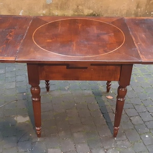 May include: A dark brown wooden table with a rectangular top, oval centre, and drop leaves. It features a drawer and four turned legs. The table is about 122 cm long when the leaves are extended.