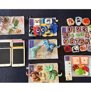 May include: A collection of educational toys, including felt sea creatures, a colorful fish, numbers, and dinosaur figures. The toys are arranged in clear pouches on a dark blue textured surface.