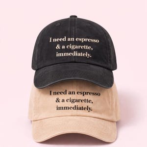 Op de afbeelding: Twee baseballpetten, één zwarte en één beige, op elkaar gestapeld. Elke pet heeft de geborduurde tekst: "I need an espresso & a cigarette, immediately."