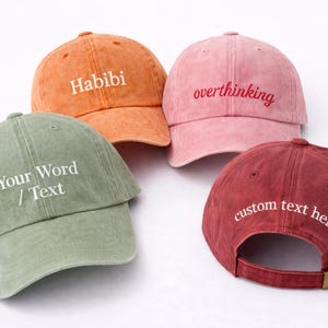 May include: Four baseball caps in shades of orange, pink, green, and burgundy. Each cap features embroidered text: "Habibi", "overthinking", "Your Word / Text", and "custom text here". The caps have adjustable straps.
