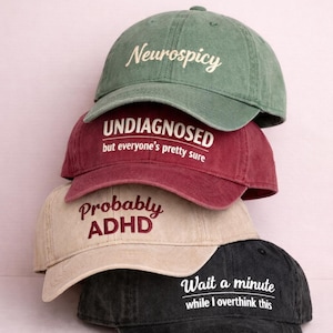 Puede incluir: Cuatro gorras de béisbol apiladas. La gorra superior es verde con la palabra "Neurospicy" bordada en blanco. La siguiente es granate con "UNDIAGNOSED but everyone's pretty sure". La tercera es beige con "Probably ADHD". La gorra inferior es negra con "Wait a minute while I overthink this".