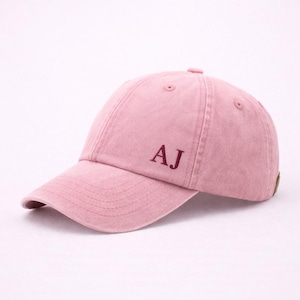 Puede incluir: Gorra de béisbol rosa empolvado con las iniciales "AJ" bordadas en un tono rojo más oscuro. La gorra tiene una visera curva y un cierre de correa de tela en la parte trasera. El sombrero está hecho de un material suave y texturizado.