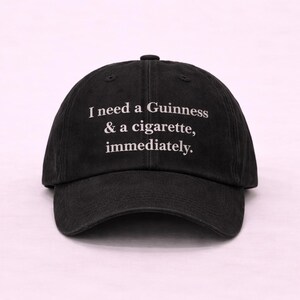 May include: Black baseball cap with the text "I need a Guinness & a cigarette, immediately." The cap has a curved brim and a fabric strap closure on the back. The text is in a white, sans-serif font.