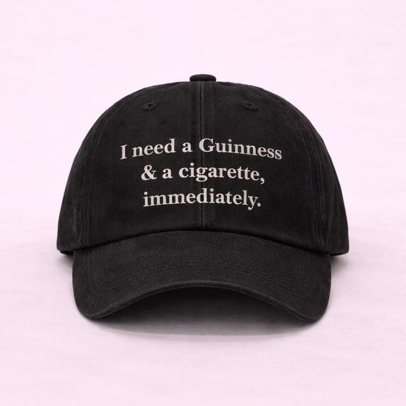 May include: Black baseball cap with the text "I need a Guinness & a cigarette, immediately." The cap has a curved brim and a fabric strap closure on the back. The text is in a white, sans-serif font.