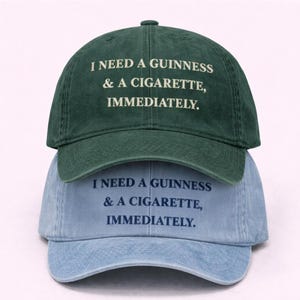 May include: Two baseball caps, one dark green and one light blue, stacked on top of each other. Each cap has the text "I NEED A GUINNESS & A CIGARETTE, IMMEDIATELY." printed in white on the green cap and dark blue on the light blue cap.