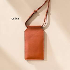 May include: A rectangular amber-colored phone pouch with a smooth finish and a matching thin adjustable shoulder strap. The pouch features a top opening and is labeled with the word Amber in black text on the upper left side of the frame.