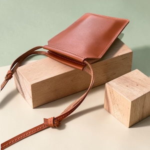 May include: A rectangular, smooth, tan leather pouch with a matching thin leather shoulder strap. The pouch features a simple, open-top design with visible stitching along the edges of the strap and the main body, presenting a clean, structured appearance.