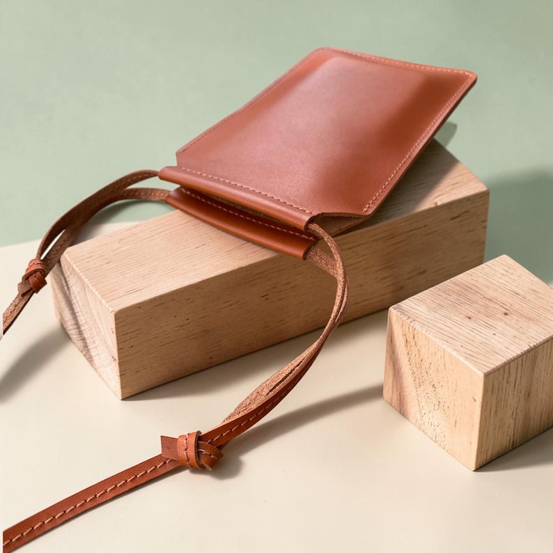 May include: A rectangular, smooth, tan leather pouch with a matching thin leather shoulder strap. The pouch features a simple, open-top design with visible stitching along the edges of the strap and the main body, presenting a clean, structured appearance.
