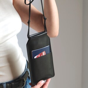 May include: A black leather crossbody phone pouch featuring a slim rectangular silhouette, a thin adjustable cord strap, and an exterior slip pocket containing a blue passport and a red credit card with visible Visa branding and a contactless symbol.
