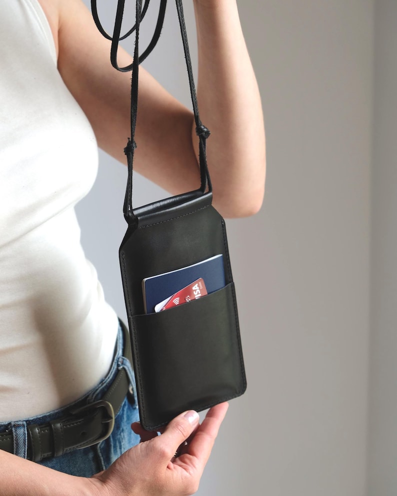 May include: A black leather crossbody phone pouch featuring a slim rectangular silhouette, a thin adjustable cord strap, and an exterior slip pocket containing a blue passport and a red credit card with visible Visa branding and a contactless symbol.