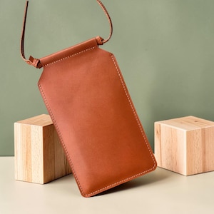 May include: A rectangular, tan leather phone pouch featuring a smooth finish, visible stitching along the edges, and a thin leather strap attached to the top. The pouch is designed to hold a mobile device securely with a simple, clean construction.