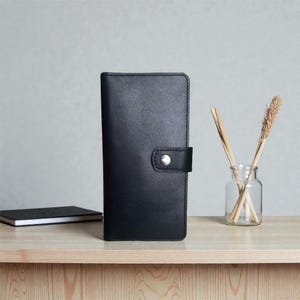 May include: A vertical black leather wallet featuring a smooth surface, rounded corners, and a single snap button closure on a small tab. The wallet is rectangular in shape and displays visible stitching along the edges and the closure tab.