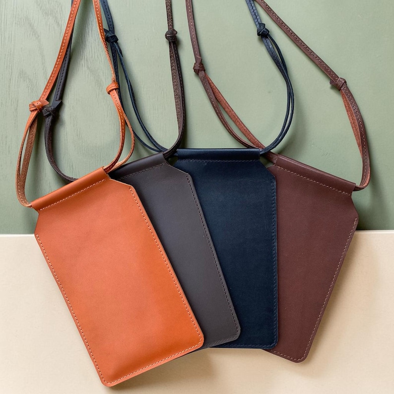 May include: Four rectangular leather pouches in tan, dark gray, navy blue, and brown, each featuring a slim adjustable leather cord strap. The pouches have a smooth finish and a simple, flat design, intended for carrying small items around the neck.