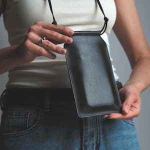Puede incluir: Una funda de cuero negro para teléfono con forma rectangular y esquinas redondeadas. La funda cuenta con una correa de cordón negro fino para llevarla alrededor del cuello o del hombro. El cuero tiene un acabado liso y mate con costuras.